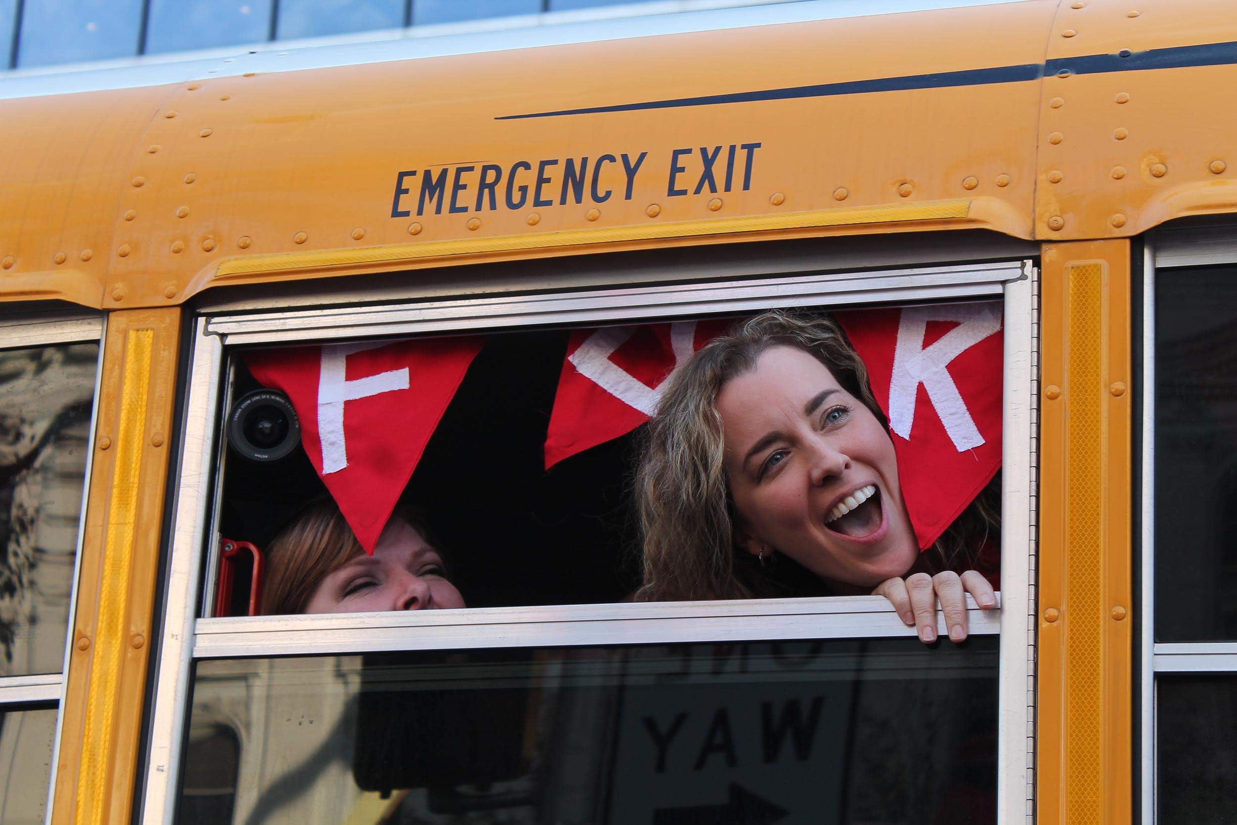 A teacher sticks their head out of the window of a school bus. A pennant flag has the letters F, T, K. They stand for 'For The Kids.'